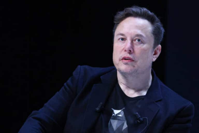 Elon Musk speaking at an event with a serious expression, addressing a massive cyberattack on X.