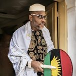 Nnamdi Kanu stands outside a doorway speaking as a security aide leans forward to listen.