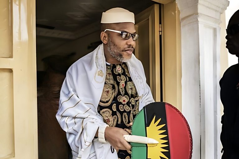 Nnamdi Kanu stands outside a doorway speaking as a security aide leans forward to listen.